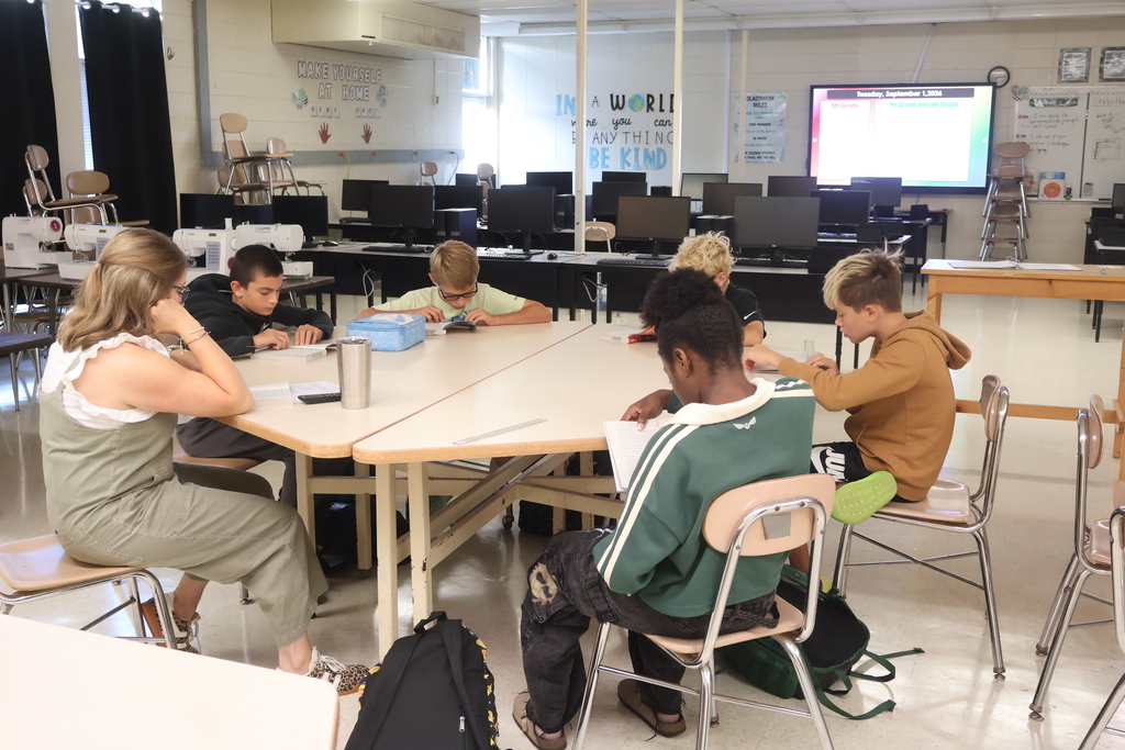 A classroom with students seated at tables, one with a backpack, and a teacher nearby. Computers and a projector are on the wall.