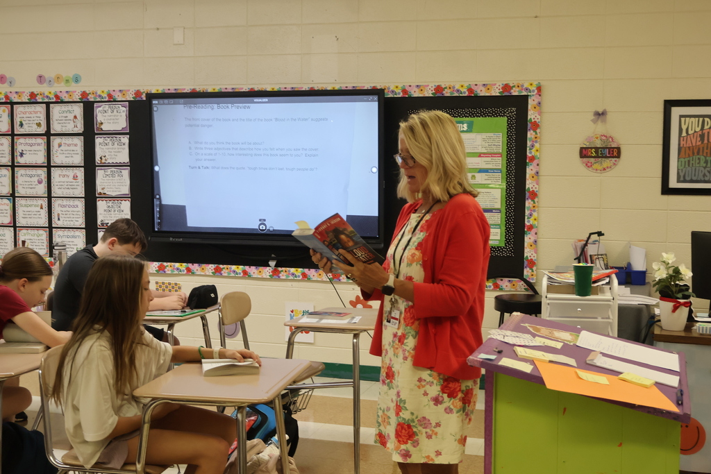 A woman in a red cardigan stands reading a book in a classroom with students seated.