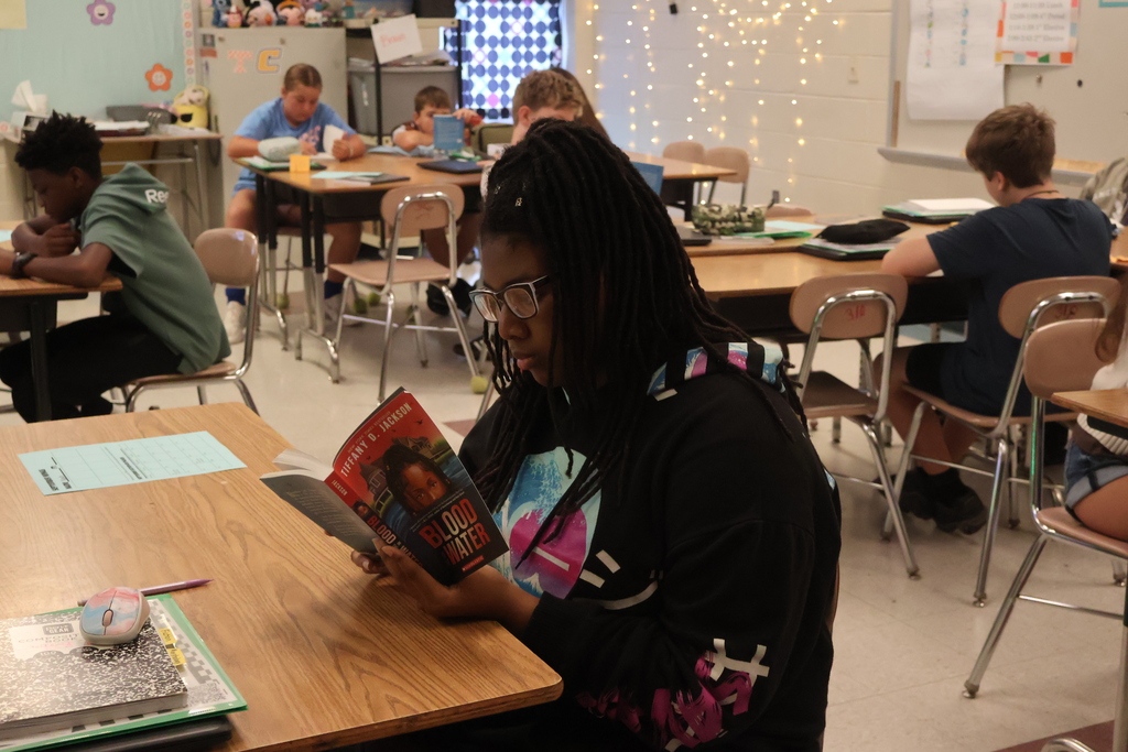 A person reading a book seated at a desk in a classroom. Other students in the background.