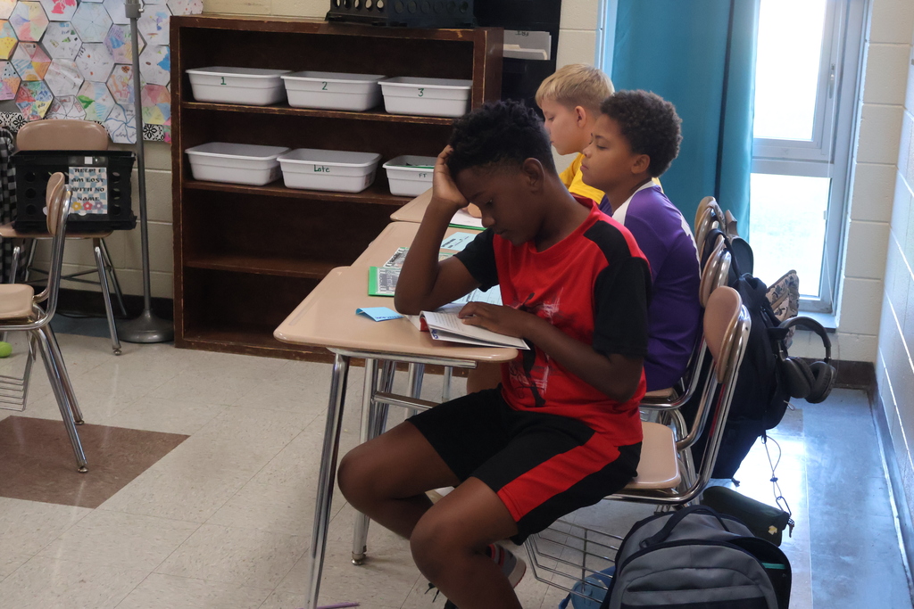 Three students are seated at desks in a classroom. One student in a red shirt rests head on hands.