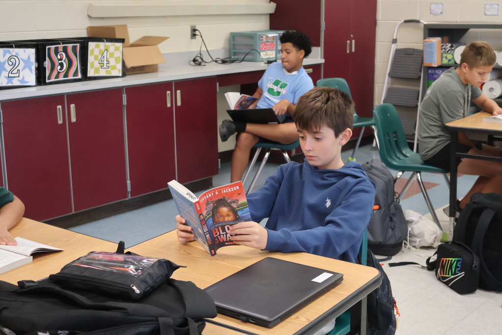 A student sits in a chair, reading a book. Other students sit at desks behind him. Backpacks and school supplies are on the desks and floor.