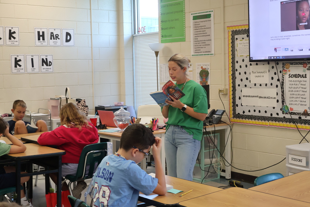 Classroom with a teacher reading aloud to students. Desks and chairs, a monitor, and bulletin board.