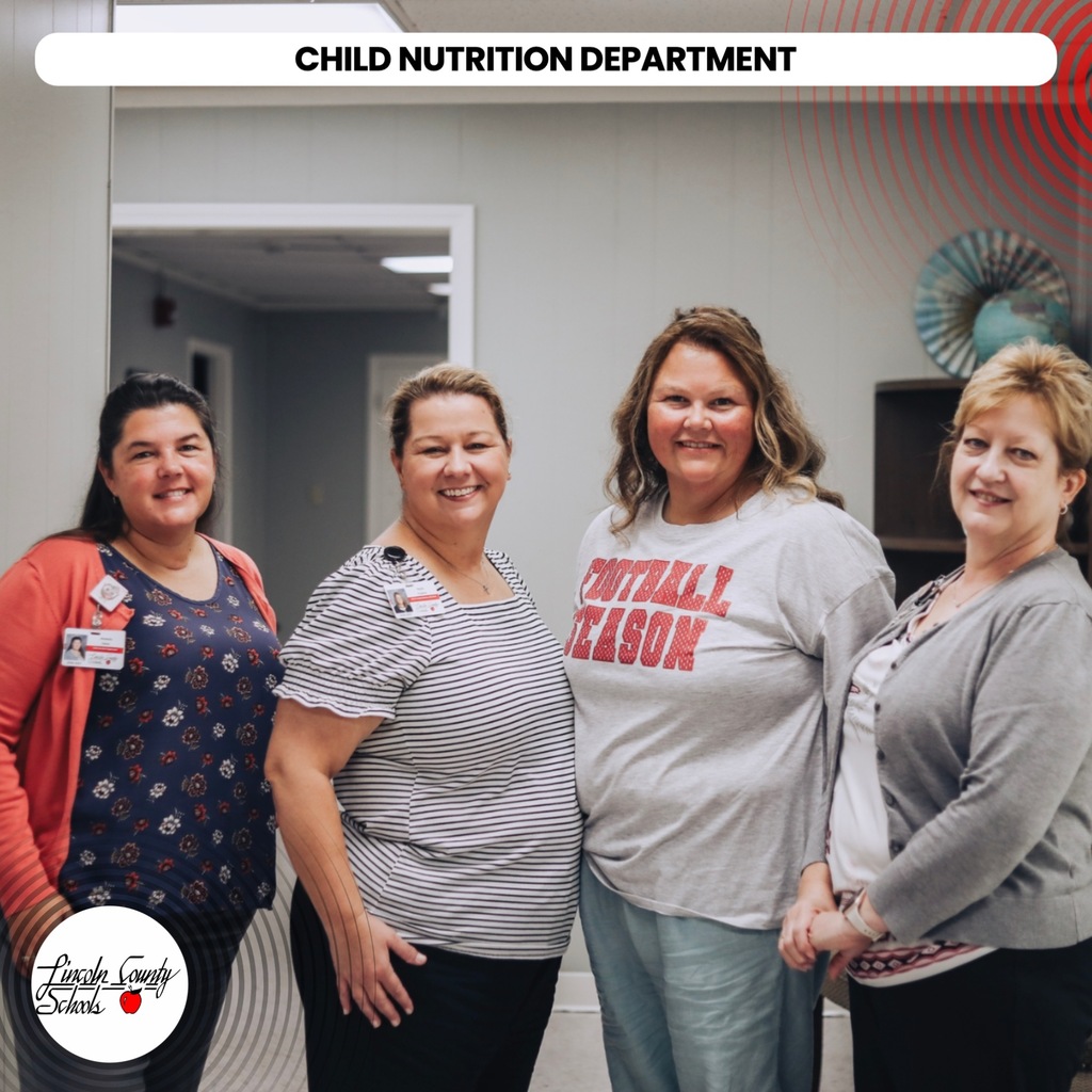 Four women in a room. They wear casual attire. The woman in the center wears a football season shirt.