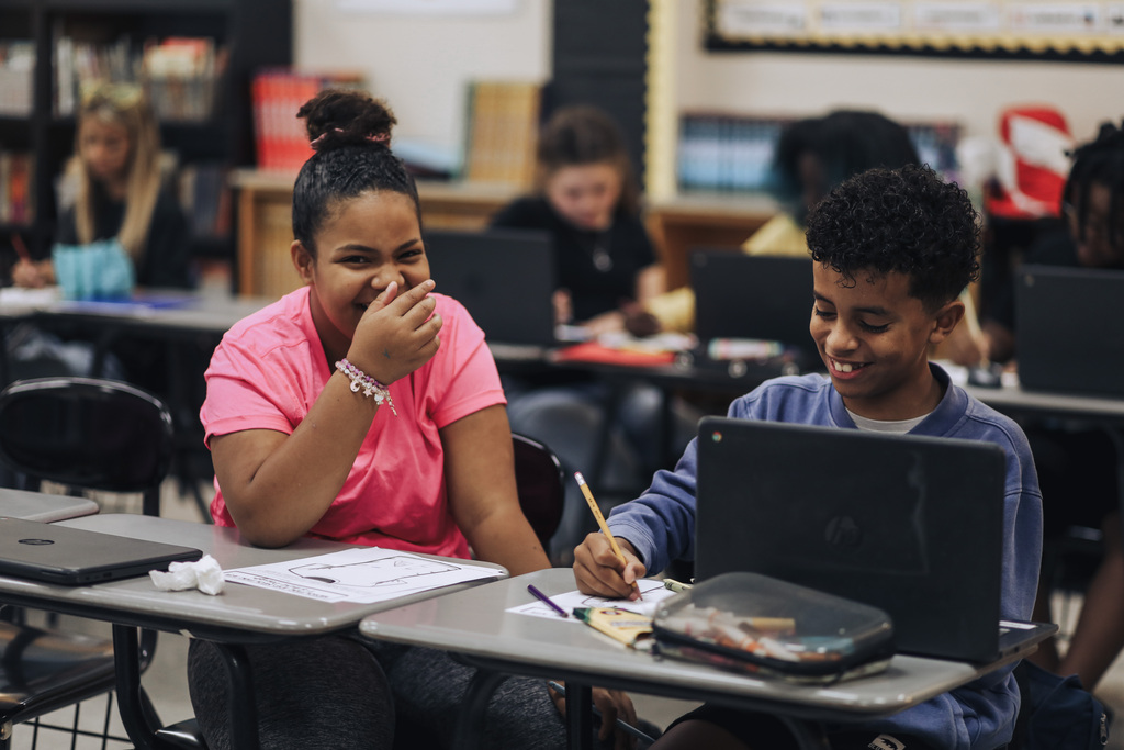 Two students, one with a pink top and one with a blue top, sit at a table with laptops, smiling.