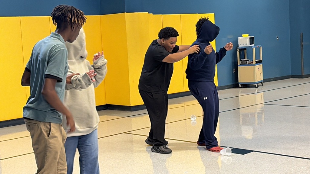 Three individuals, one in a hoodie, stand in a gym with yellow walls. One person assists another in arm movements.