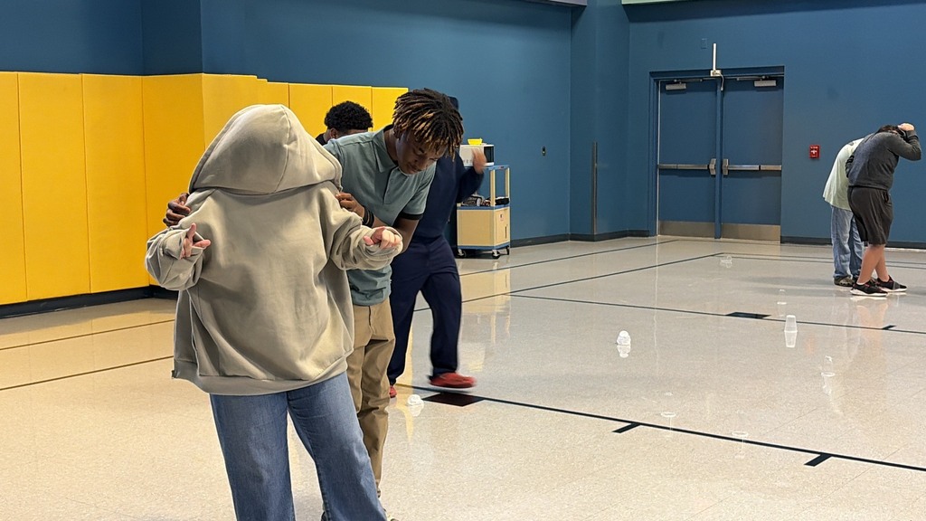 A group of people in a gym-like setting with yellow and blue walls. Some engage in a game.