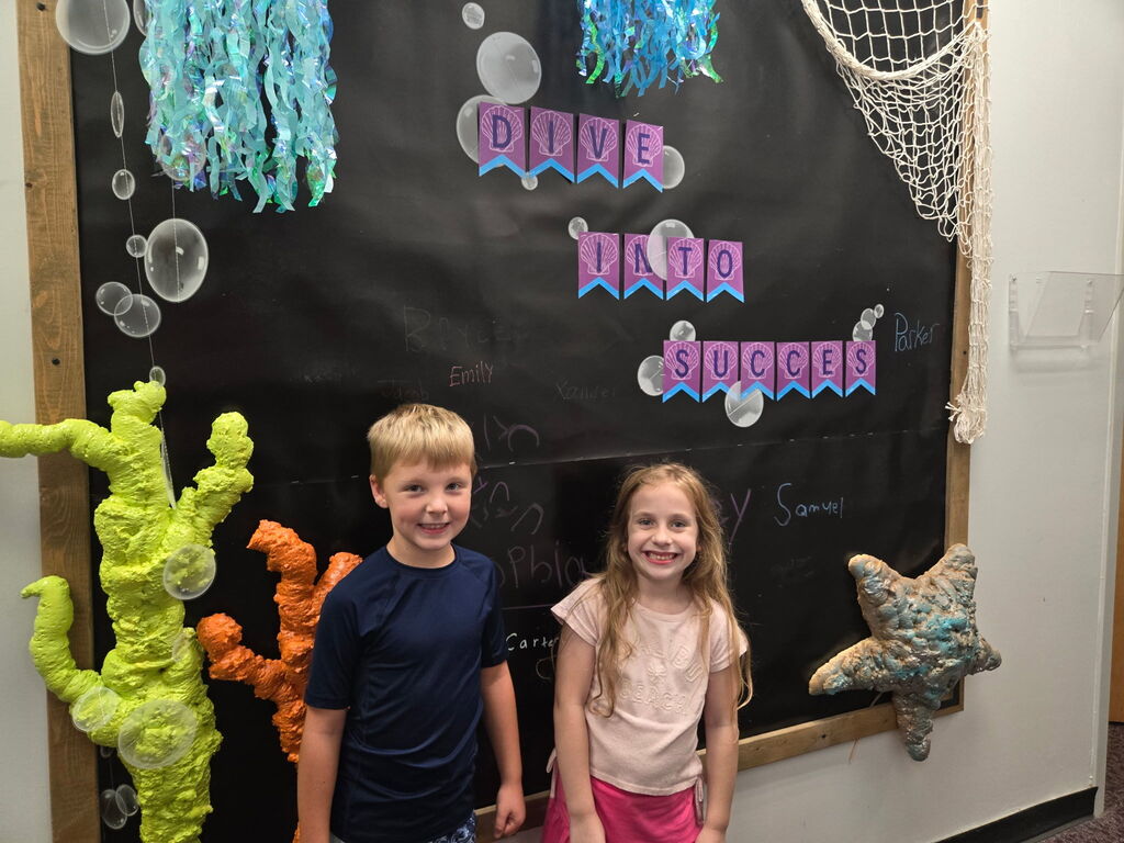 Two children, a boy and a girl, pose next to a decorated bulletin board featuring a black background, coral, bubbles, and a net.