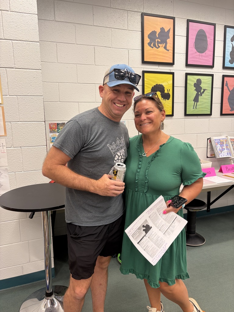 Man and woman smiling, standing in a room with colorful artwork and tables; he holds a can.