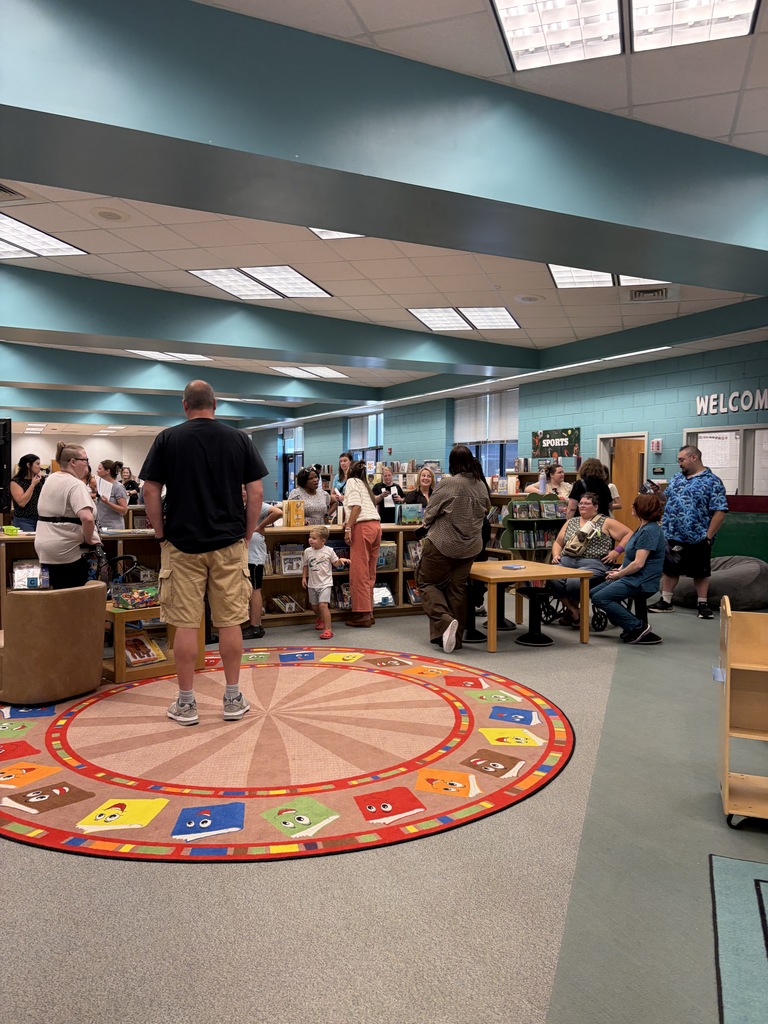 Indoor library with blue walls. People are gathered around tables, with one man standing on a colorful rug.