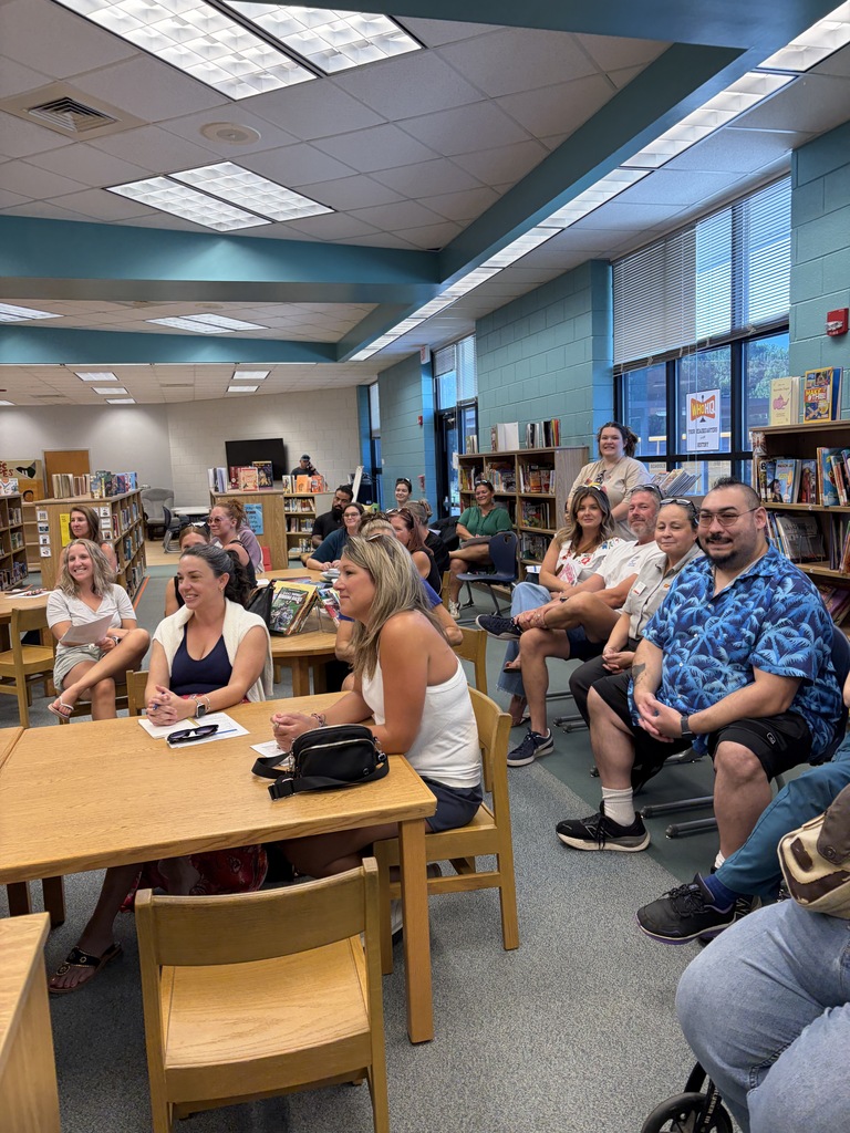 A group of people sit in chairs around tables in a library, some standing. The room has bookshelves, overhead lighting, and windows.