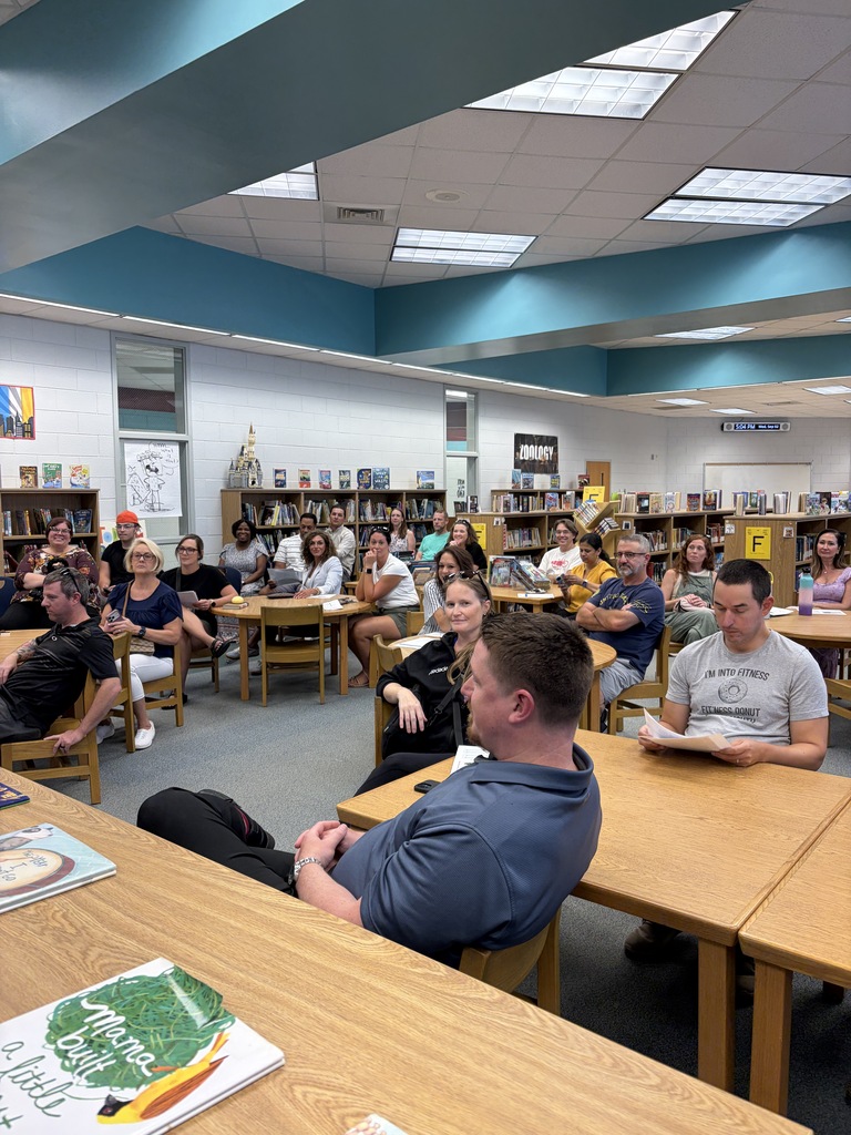 Many people seated around tables in a library, some reading, with books and desks visible.