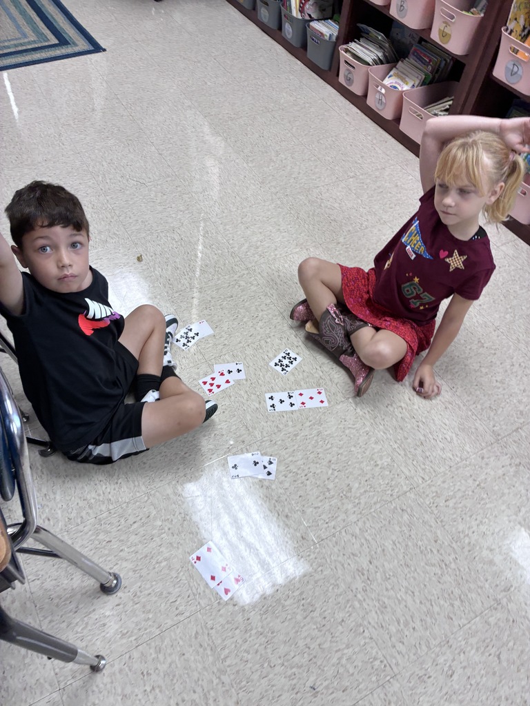 Two children sit on a classroom floor, engaged in a card game. A boy wears a black shirt, while a girl wears a red shirt. Cards are scattered around them.