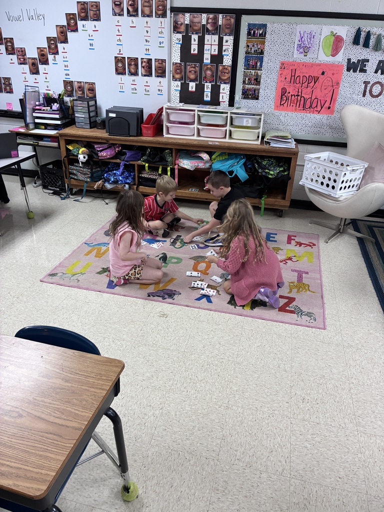Four children play with letters on a mat in a classroom. A shelf is behind them.