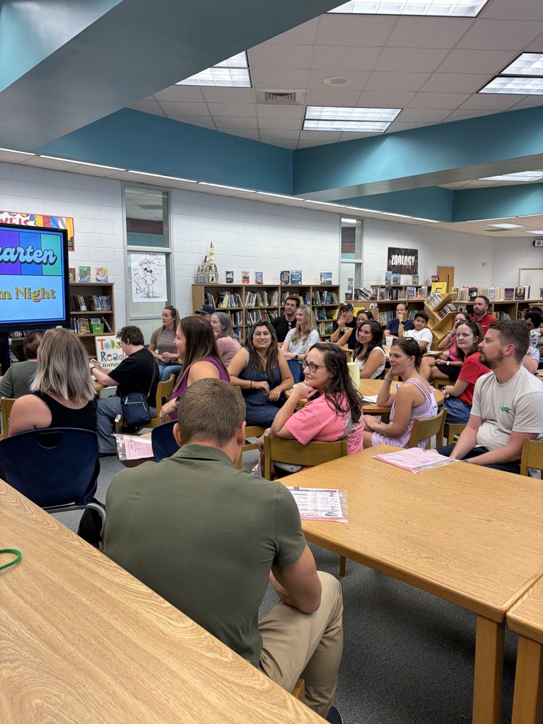 Many people sit at tables in a library. A large screen shows an image. Shelves filled with books are in the background.