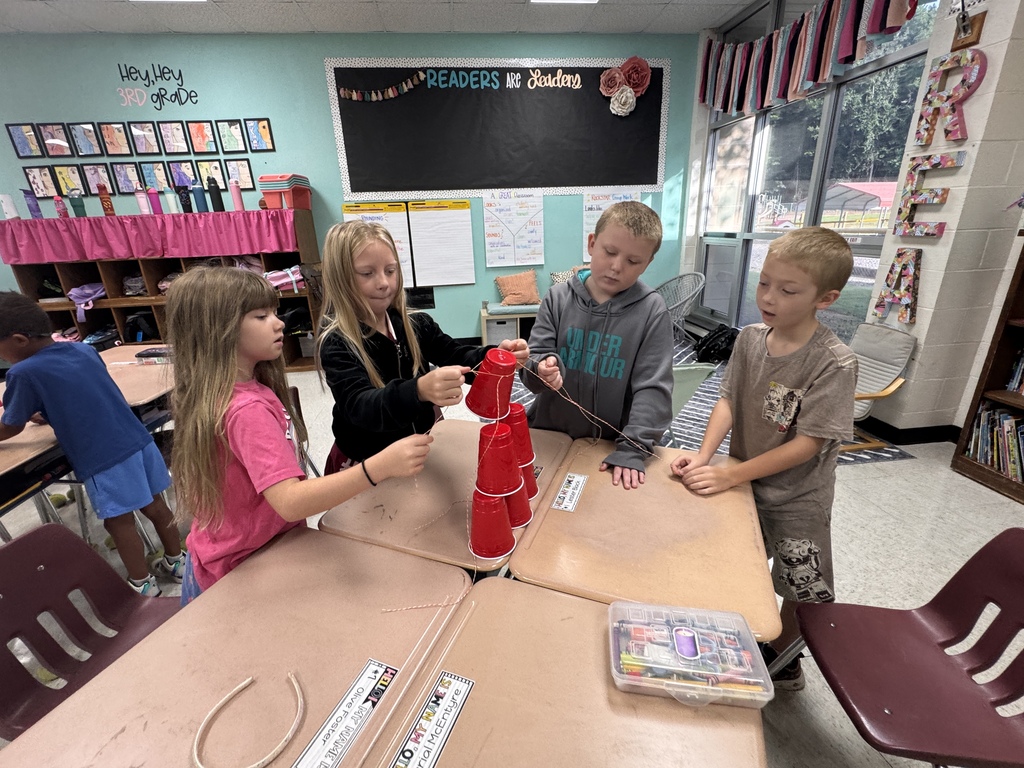 Four third-grade students gathered around classroom desks collaborating on a STEM activity, using strings tied to a rubber band to build a tower out of stacked red plastic cups.