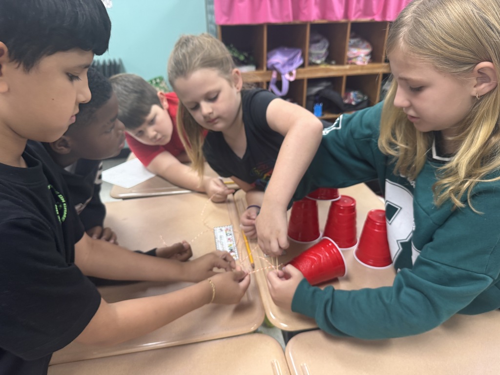 Five elementary students leaned over classroom desks working together on a hands-on STEM project, carefully manipulating strings and rubber bands to move and stack red plastic cups.