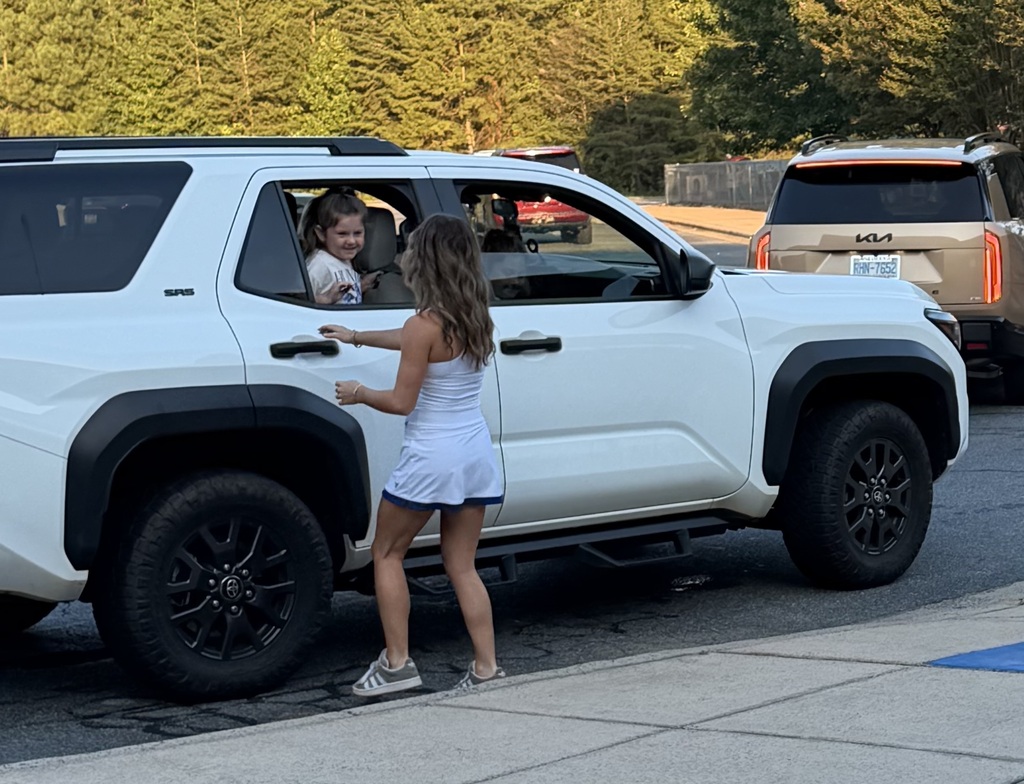 Person stands by white SUV, assisting child in vehicle. Trees and other vehicles are in the background.