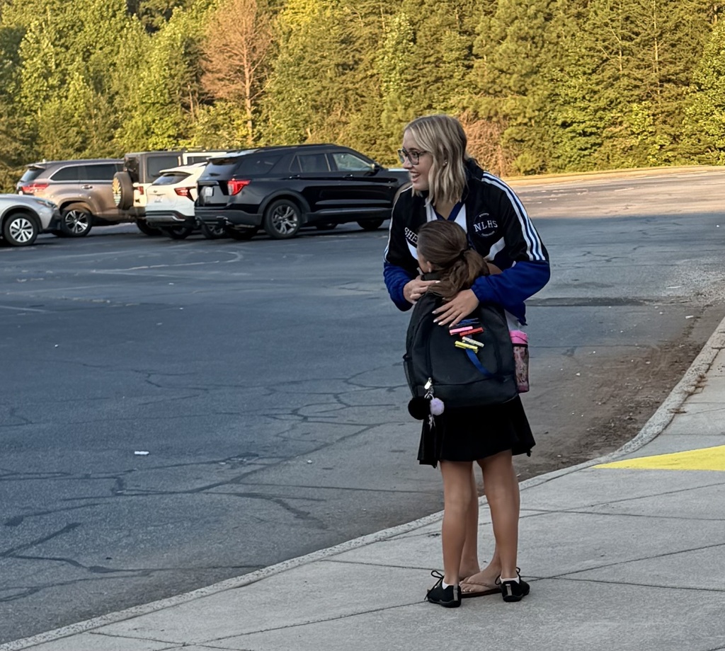 A woman and child stand on a sidewalk, facing parked cars and greenery. The woman wears glasses and a jacket.