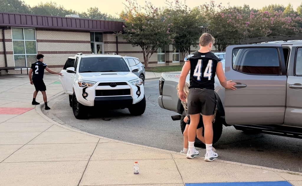 Two men in sports uniforms near vehicles, one holding a pickup truck door. A building with glass windows in the background.