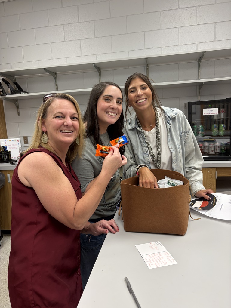 Three women stand by a table, one holding a candy bar. A brown bag is on the table.