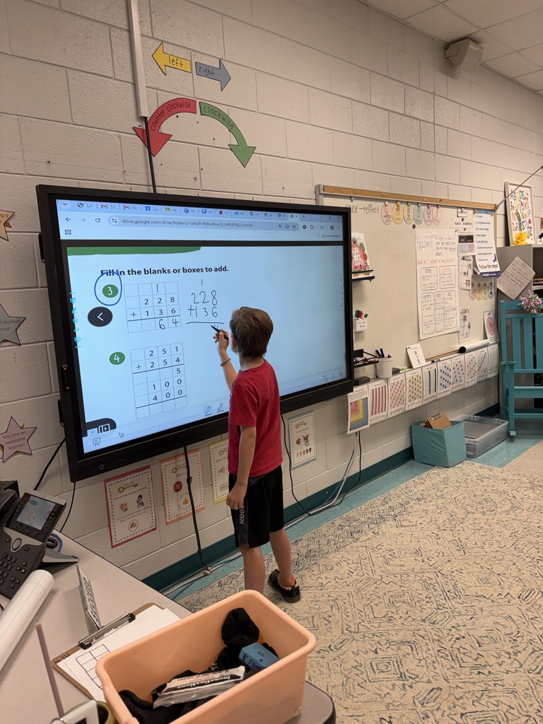 A boy in a red shirt uses a marker on a classroom smart board with a math problem.