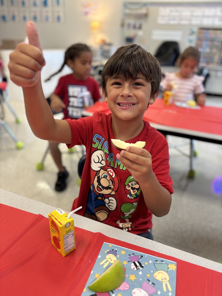 A boy with a thumbs up holds a slice of apple. Behind him, other children sit at tables.