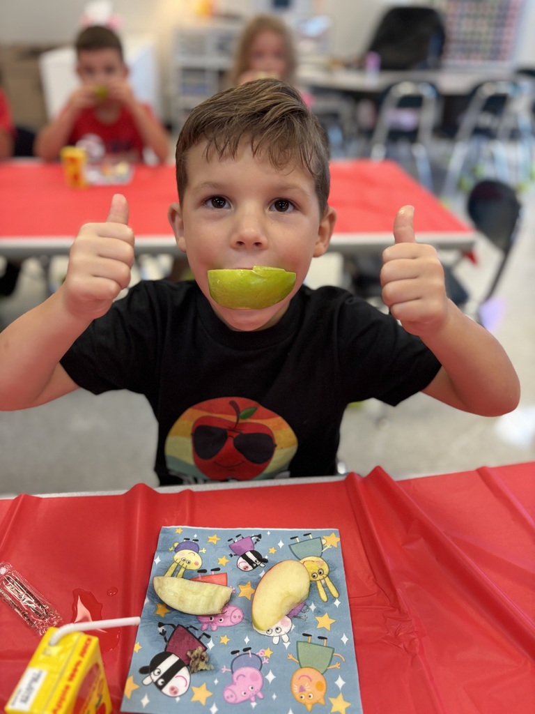 A boy sits at a table eating an apple slice, giving a thumbs-up, with a red tablecloth.
