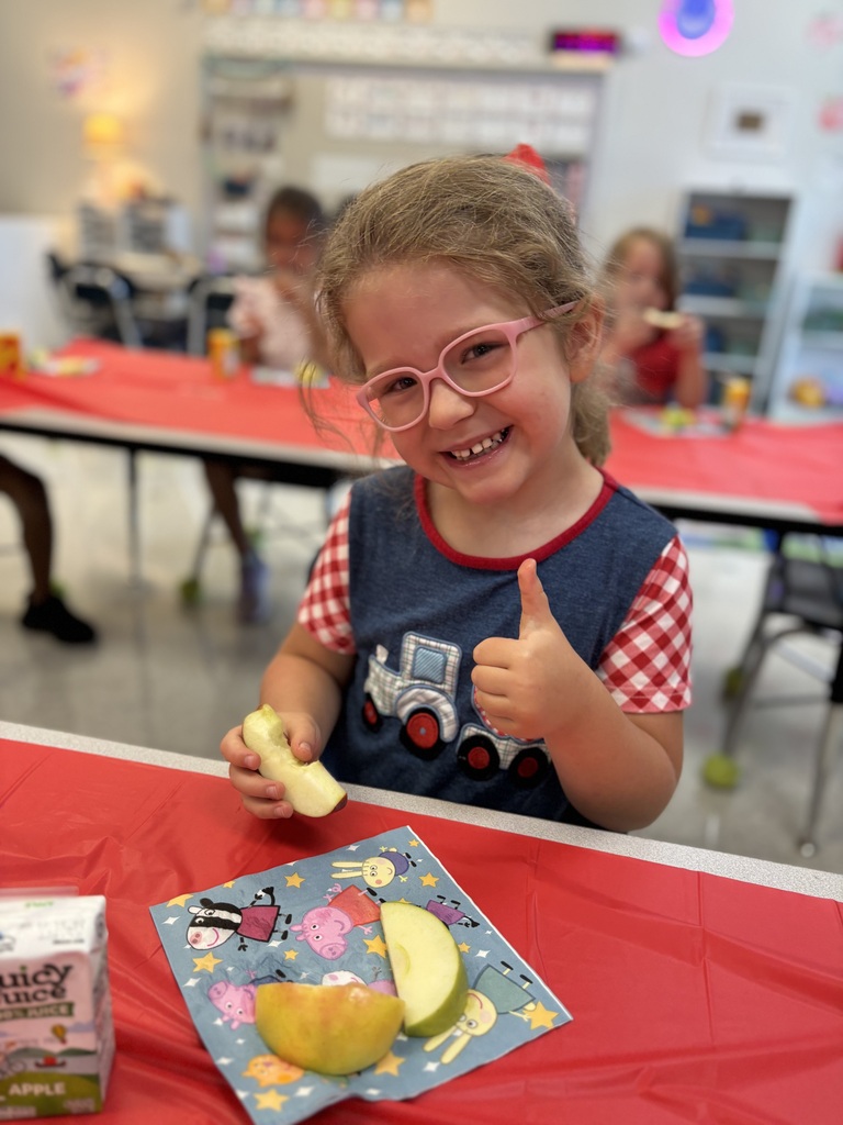 A young girl in glasses sits at a table, smiling and giving a thumbs up while holding an apple slice.