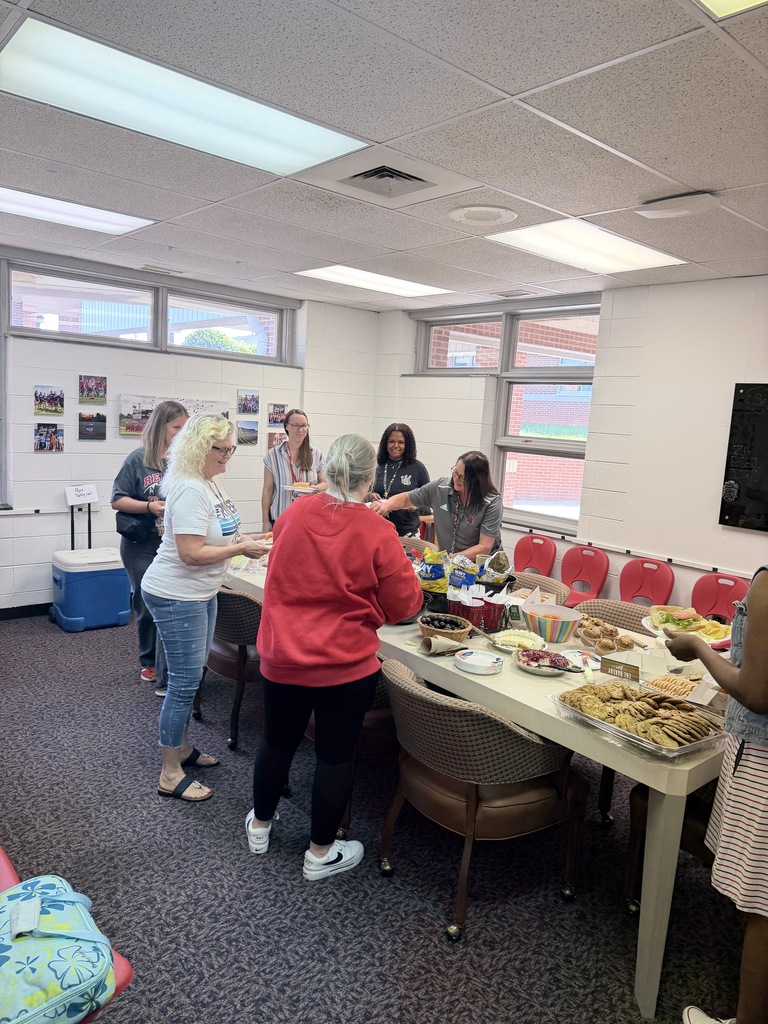 A group of people in a room, standing around a table with various foods and drinks.