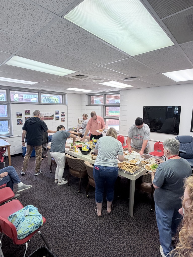 Several people stand and walk around a table with food. A television is mounted on the wall.