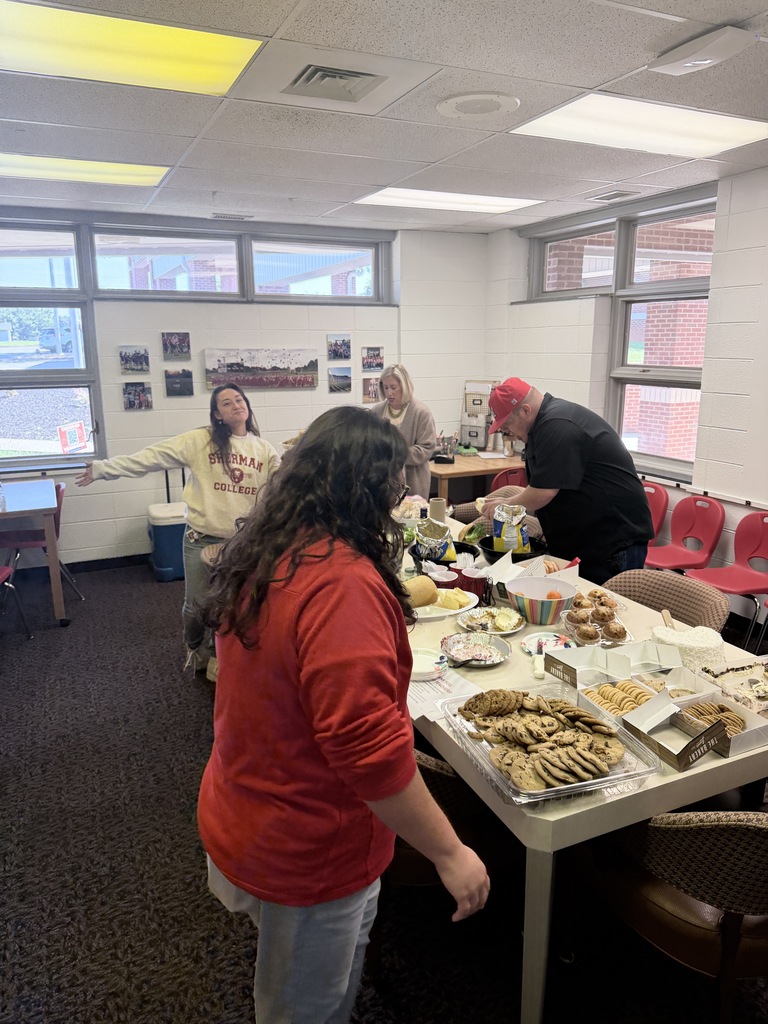 A room with several people. A woman stands before a table filled with various food items.