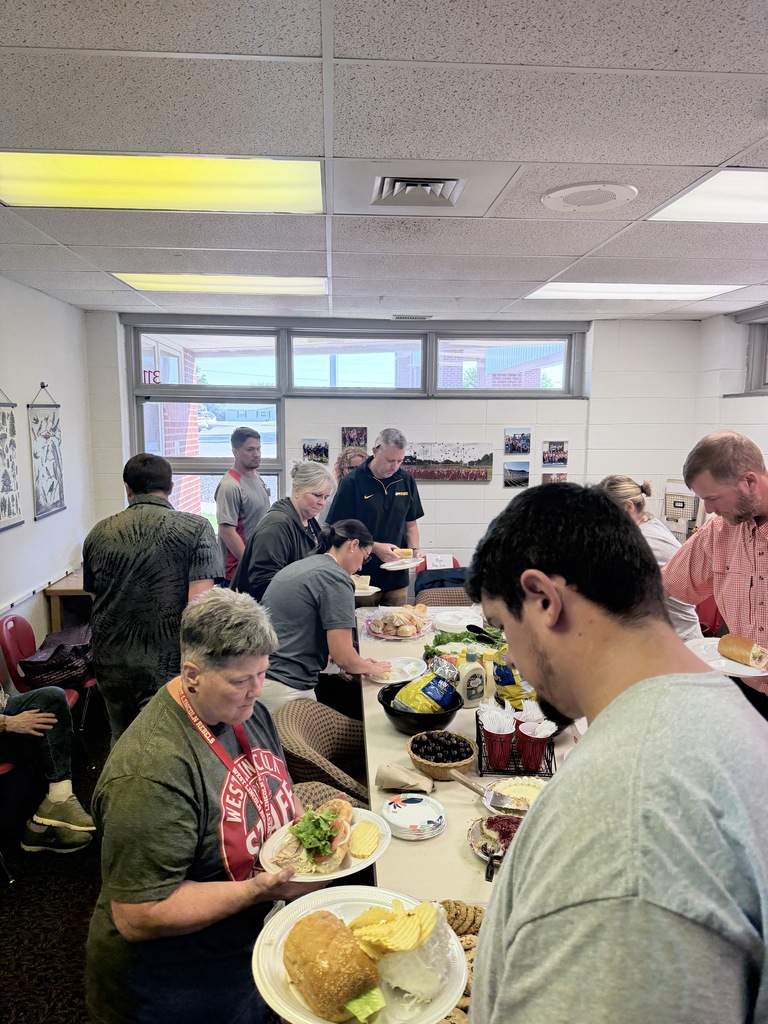 People gather at a long table, picking up plates of food. A woman wears an apron.