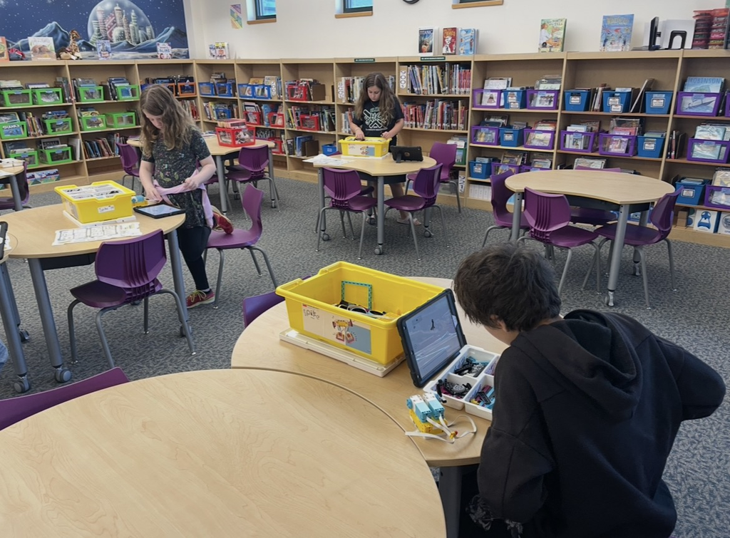 Children in a library setting use tablets on round tables. One child works at a table with a yellow box.