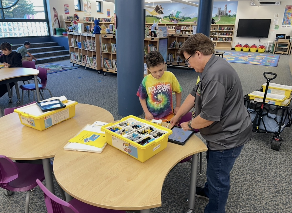 A man and a child in a library are using a table with a tablet and yellow bins.