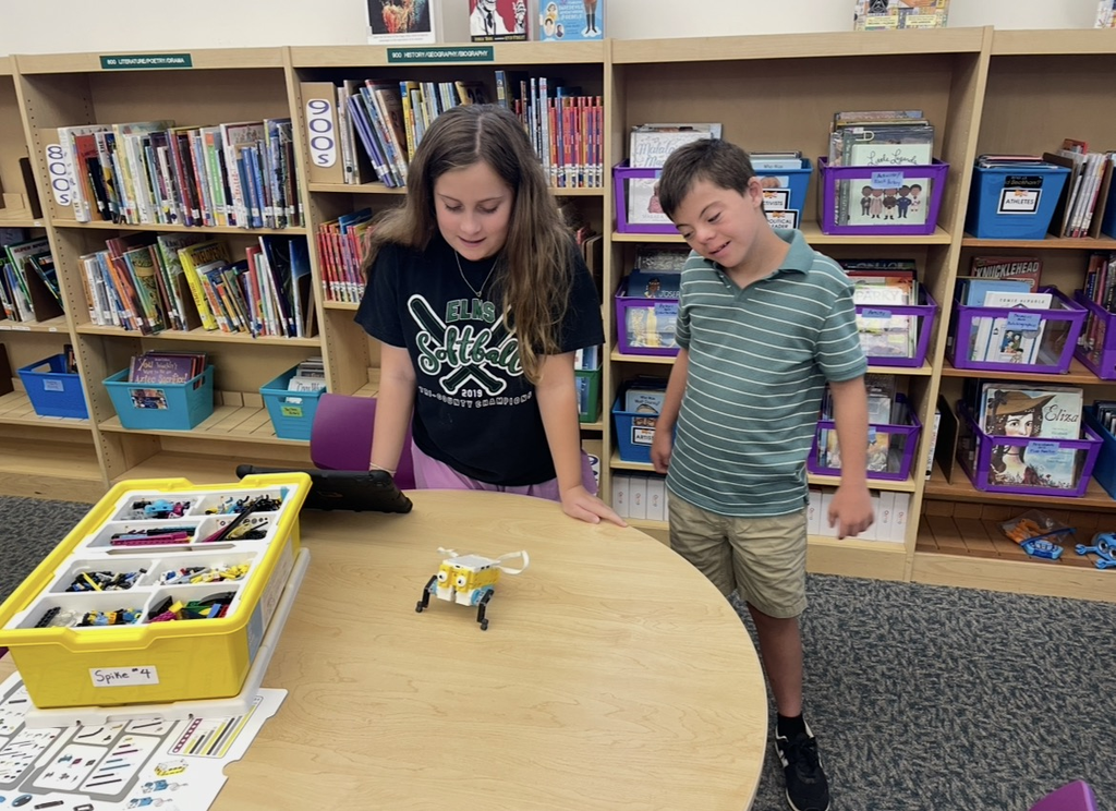 Two children are standing at a table with a robot in front of them, in a library.