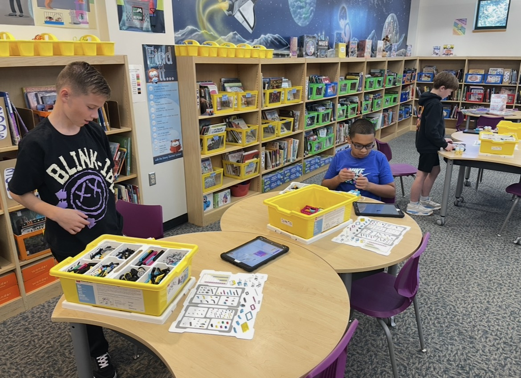 Three children in a library with a large bookshelf behind them, one standing at a table with a tablet.