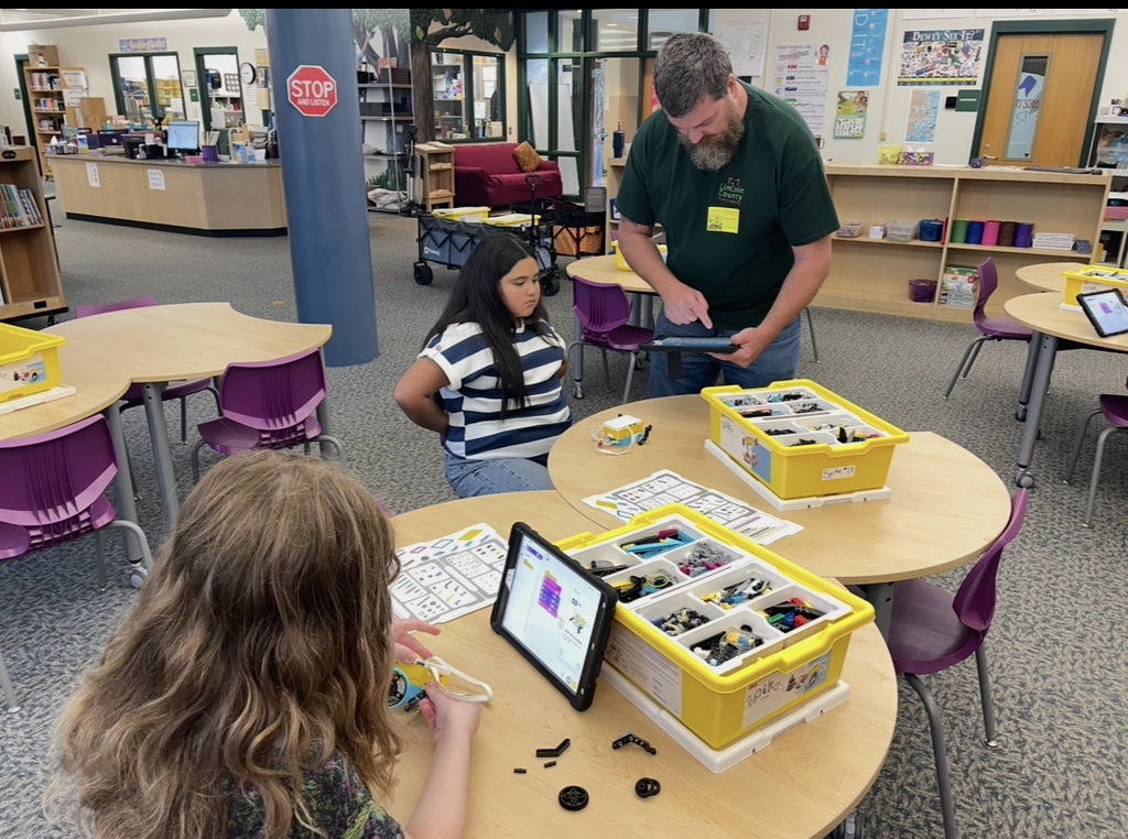 A teacher helps two students in a classroom with a table of lego parts.