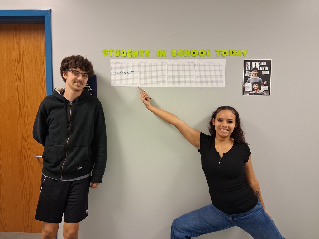 Two people standing by a wall. One points to a sign that reads "Students in School Today."