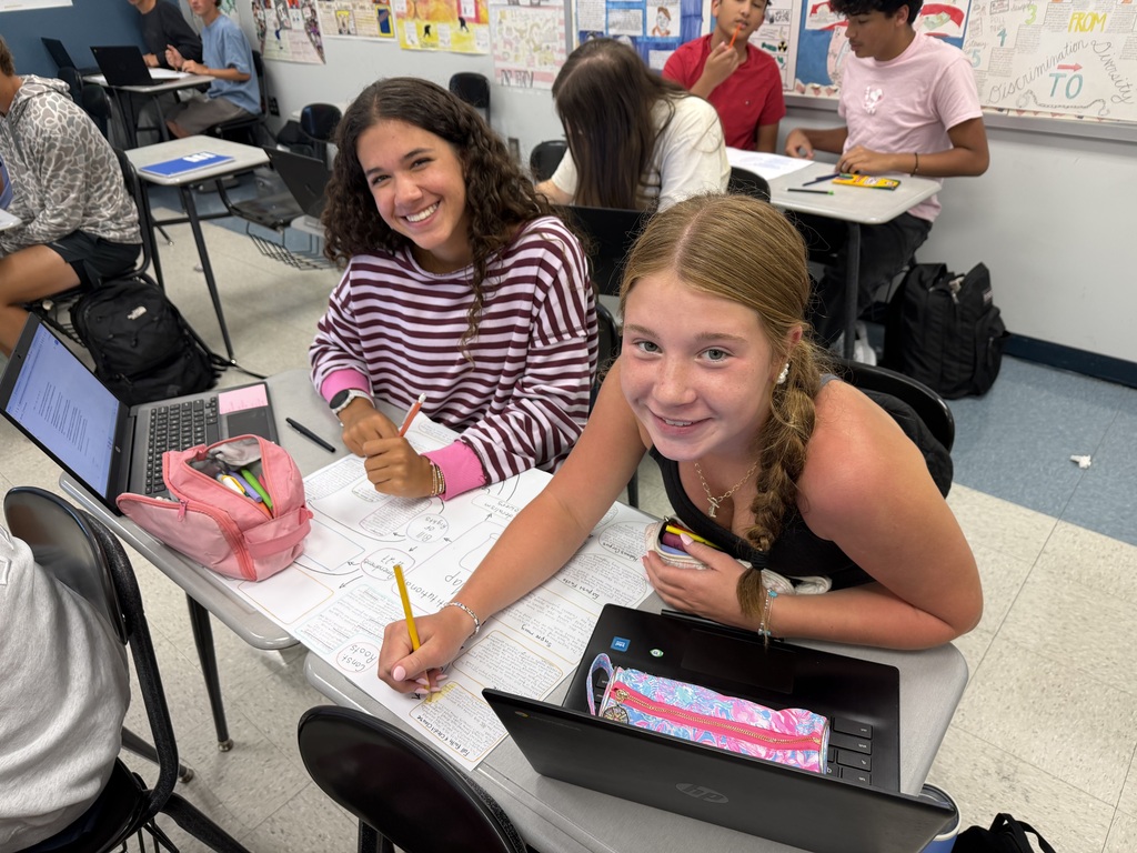 Two girls seated at a desk, one writing while the other smiles, with laptops, notebooks, and pens around them.