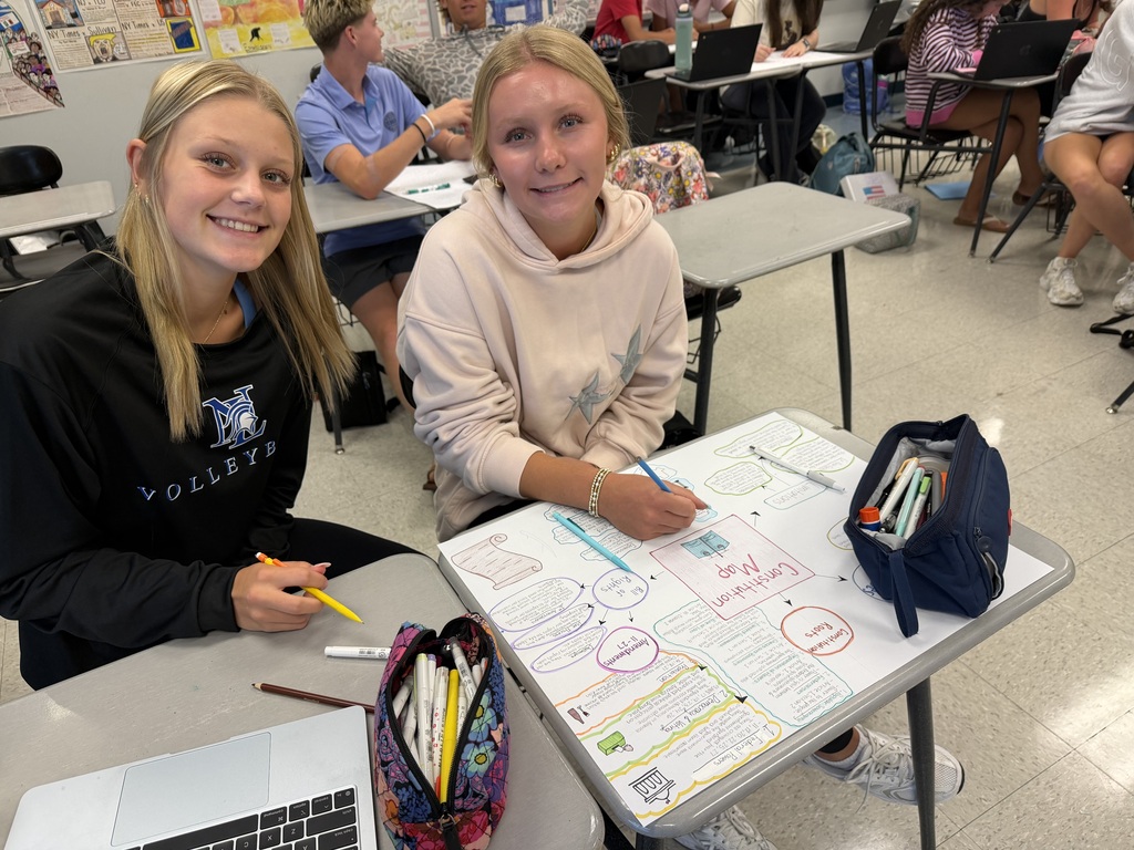 Two young women sitting at a table with laptops, pens, and paper. Other students are seated at tables in the background.