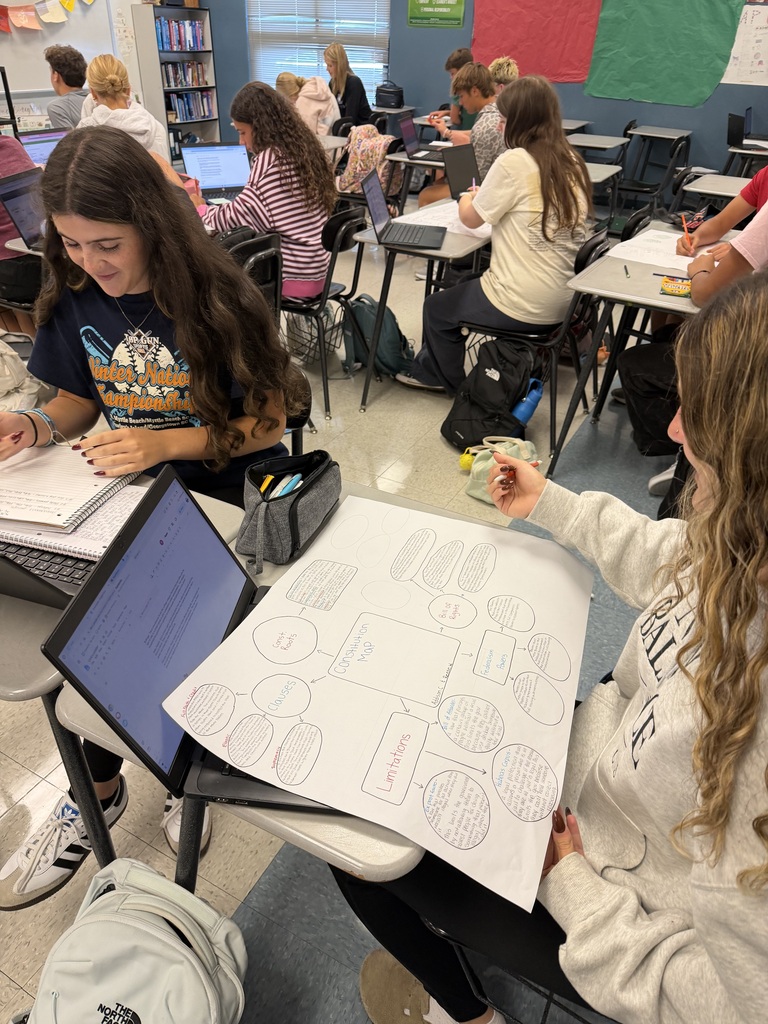 Students in a classroom seated at desks, using laptops. One student holds a large printed sheet, and another has a backpack nearby.