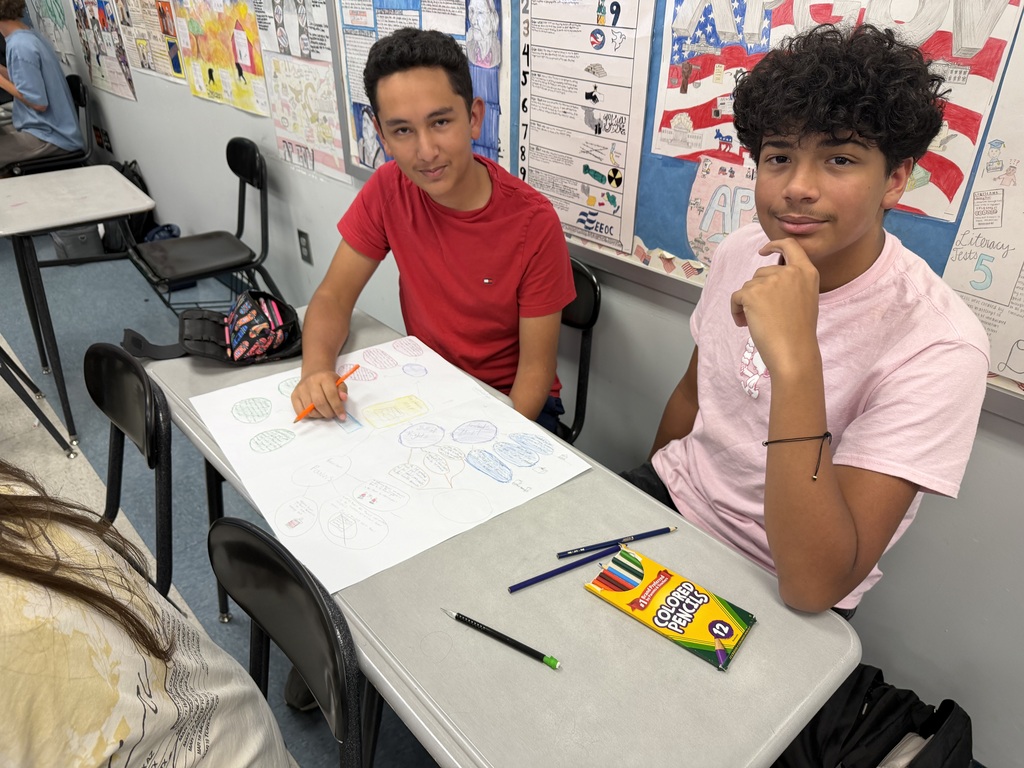 Two boys sit at a table in a classroom with a large paper and colored pencils.