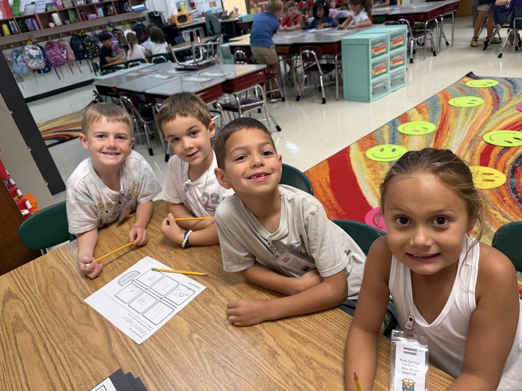 Four children, three boys and one girl, sitting at a table with papers and pencils in a classroom.