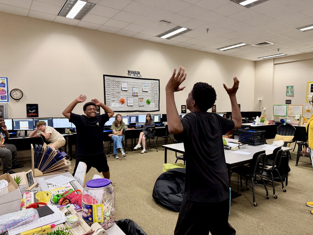 Kids in a classroom with desks, tables, and chairs. One stands with raised arms in the center.