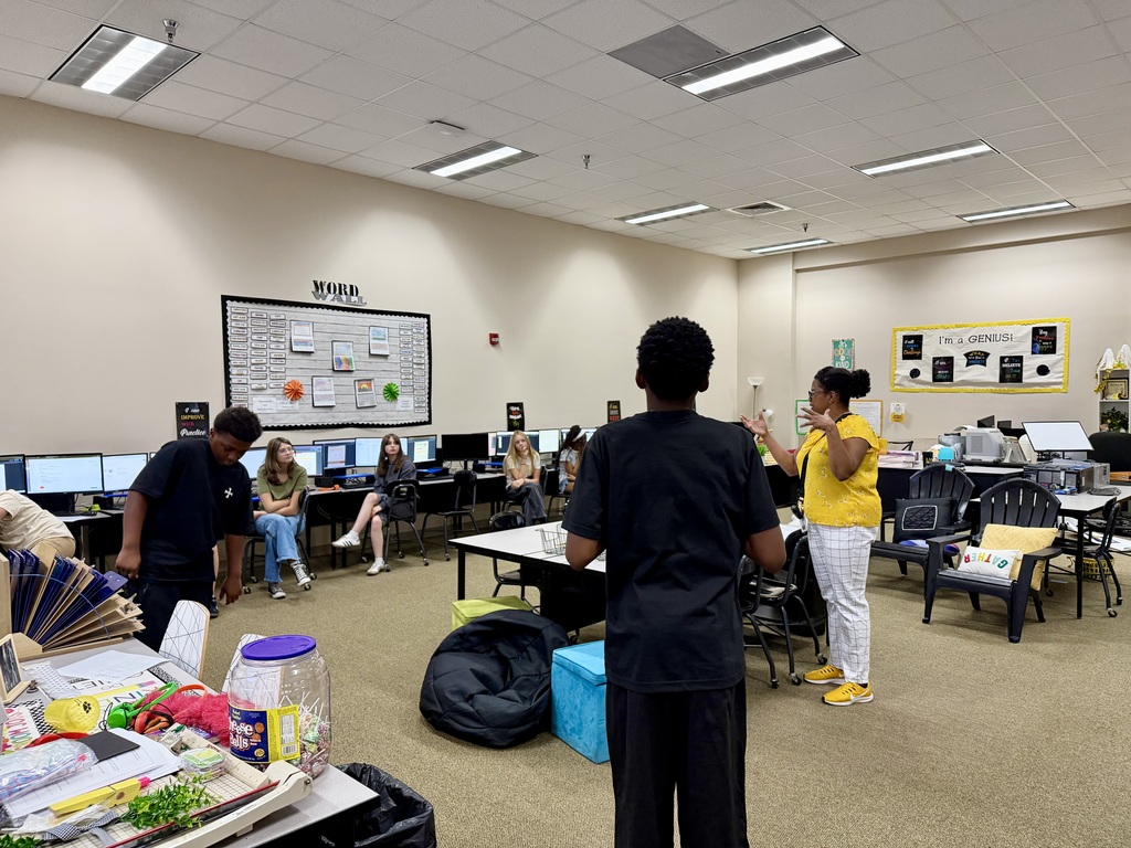 A room with people standing and sitting. One person is speaking. Tables have computers, bags, and other items.