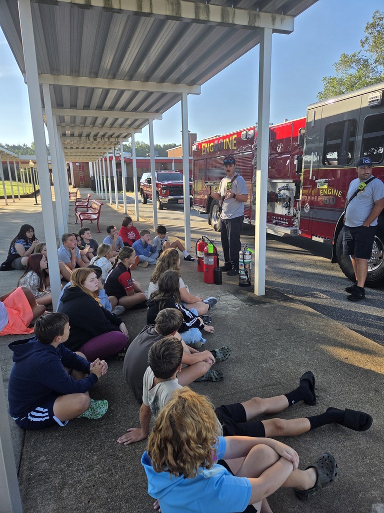 Several people, including children, sit on the ground while a firefighter stands behind them, explaining something.