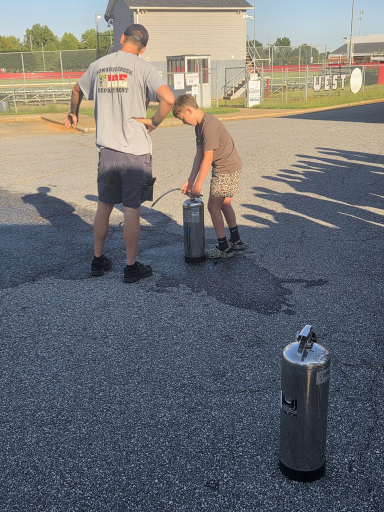 An adult and a child stand near two kegs, with one child using a hose. The background shows a building.