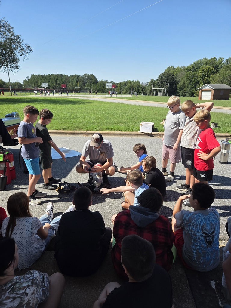 Group of children and adults seated and standing in a circle, on a paved area with a grassy field in the background.
