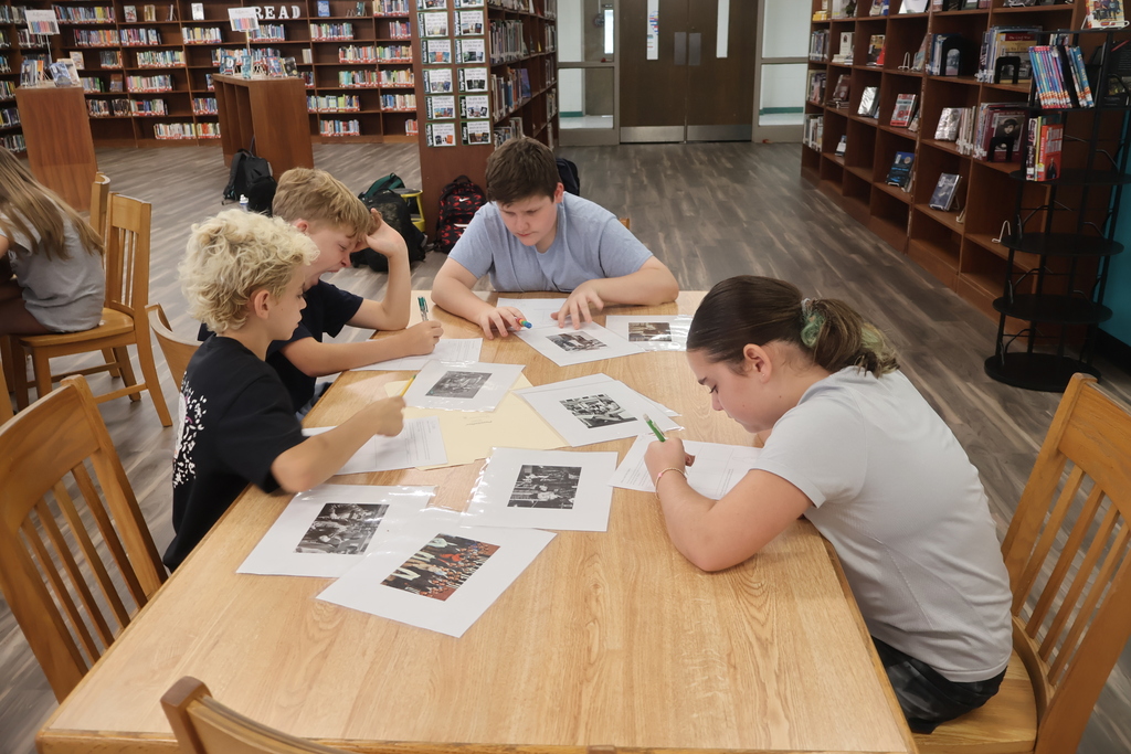 Five children sit around a library table, working on papers with printed pictures. Bookshelves are in the background.