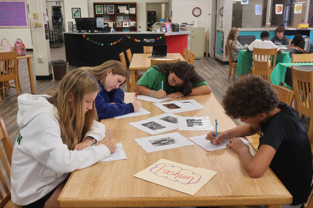 Four students sit at a table, writing on papers. A table has papers and a sign saying Fashion.